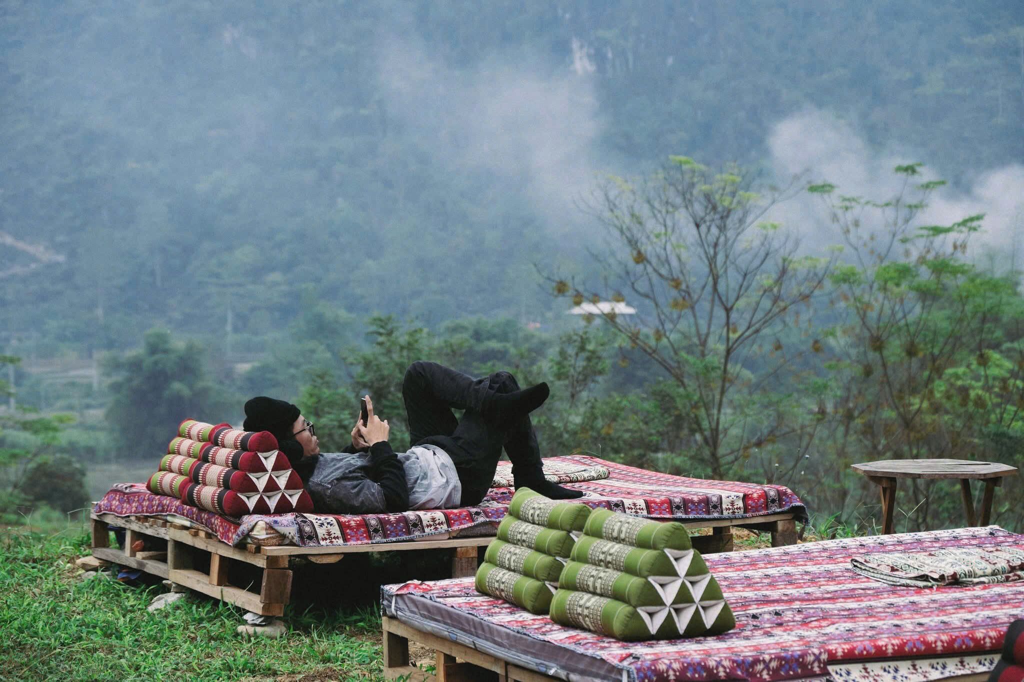 A person lying on a bed with a patterned blanket, surrounded by neatly stacked pillows and a small table.