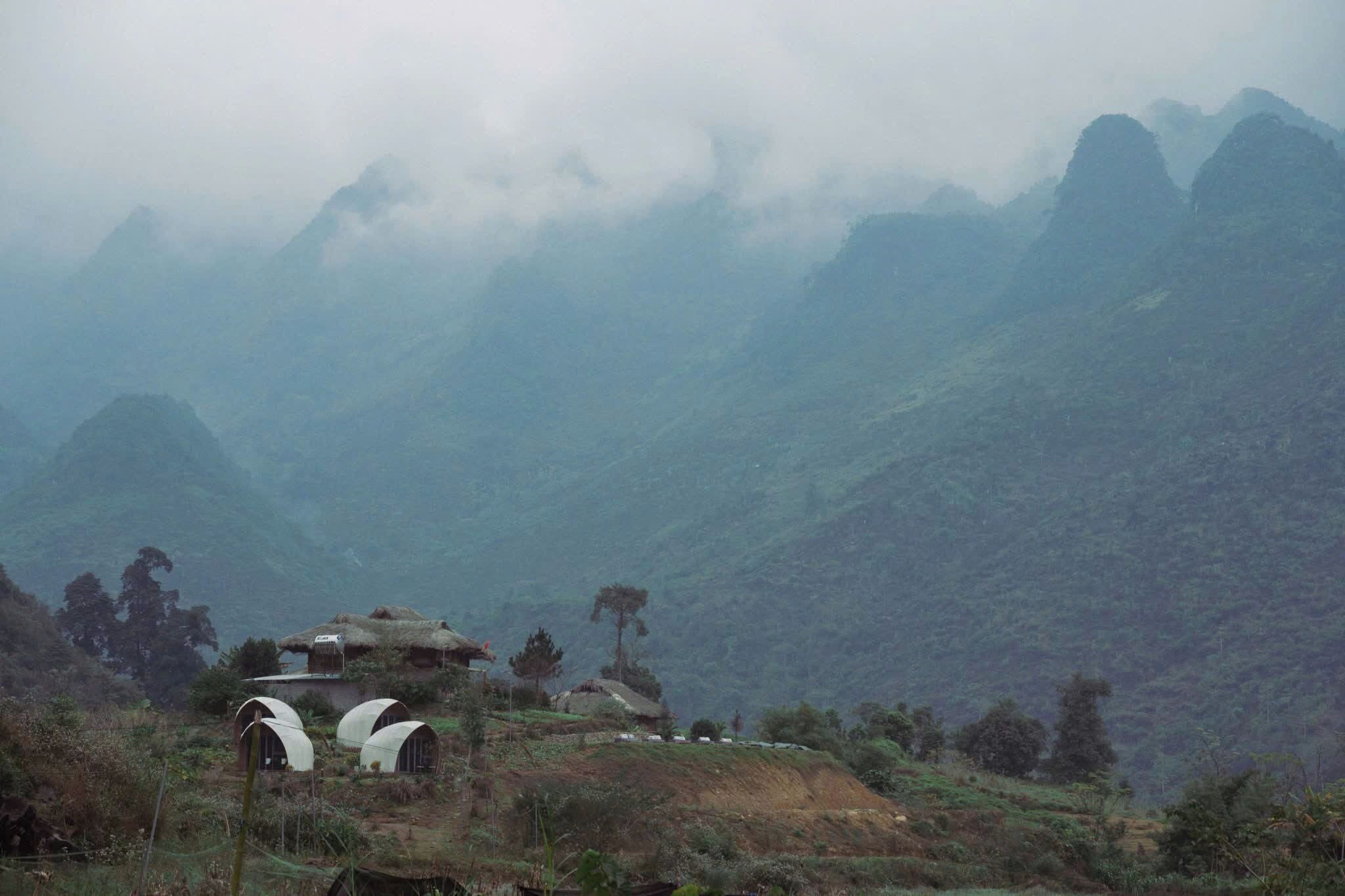 A mountainous landscape with traditional thatched-roof huts and a few modern structures, surrounded by dense forest and mist.