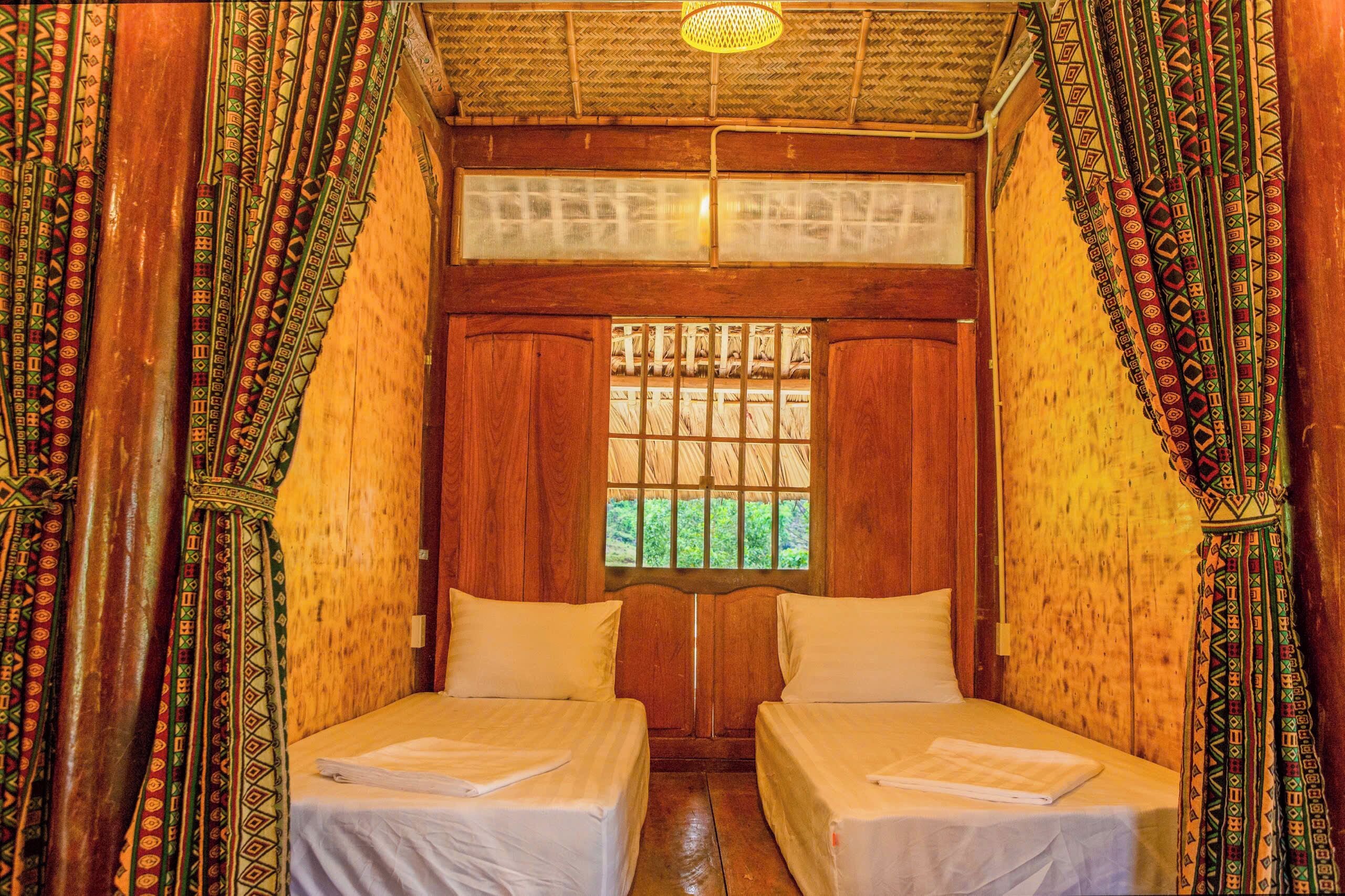 Una habitación con dos camas, paredes de madera, una ventana con barrotes y cortinas estampadas.