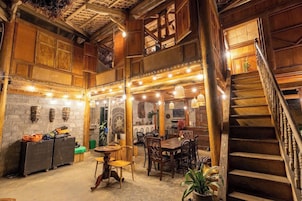 Interior rústico com escada de madeira, área de jantar com mesas e cadeiras, e parede de tijolos com detalhes decorativos.