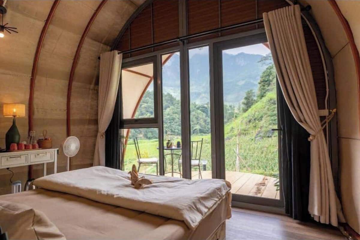 A bedroom with a bed, a desk, a fan, and a view of a mountainous landscape.