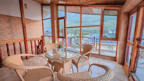 A cozy room with a glass table surrounded by wicker chairs, large windows offering a view of a mountainous landscape, and a wooden staircase. - Hotel Jubilant Manali (Manali)