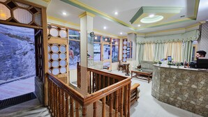 A hotel lobby with a wooden reception desk, a stone accent wall, and a view of the outside through large windows. - Hotel Jubilant Manali (Manali)
