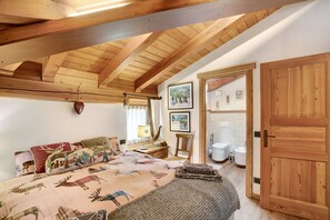 Een slaapkamer met een houten plafond, een bed met een sprei met daop afbeelding van een eland, een nachtkastje met een lamp, en een deur naar een badkamer.