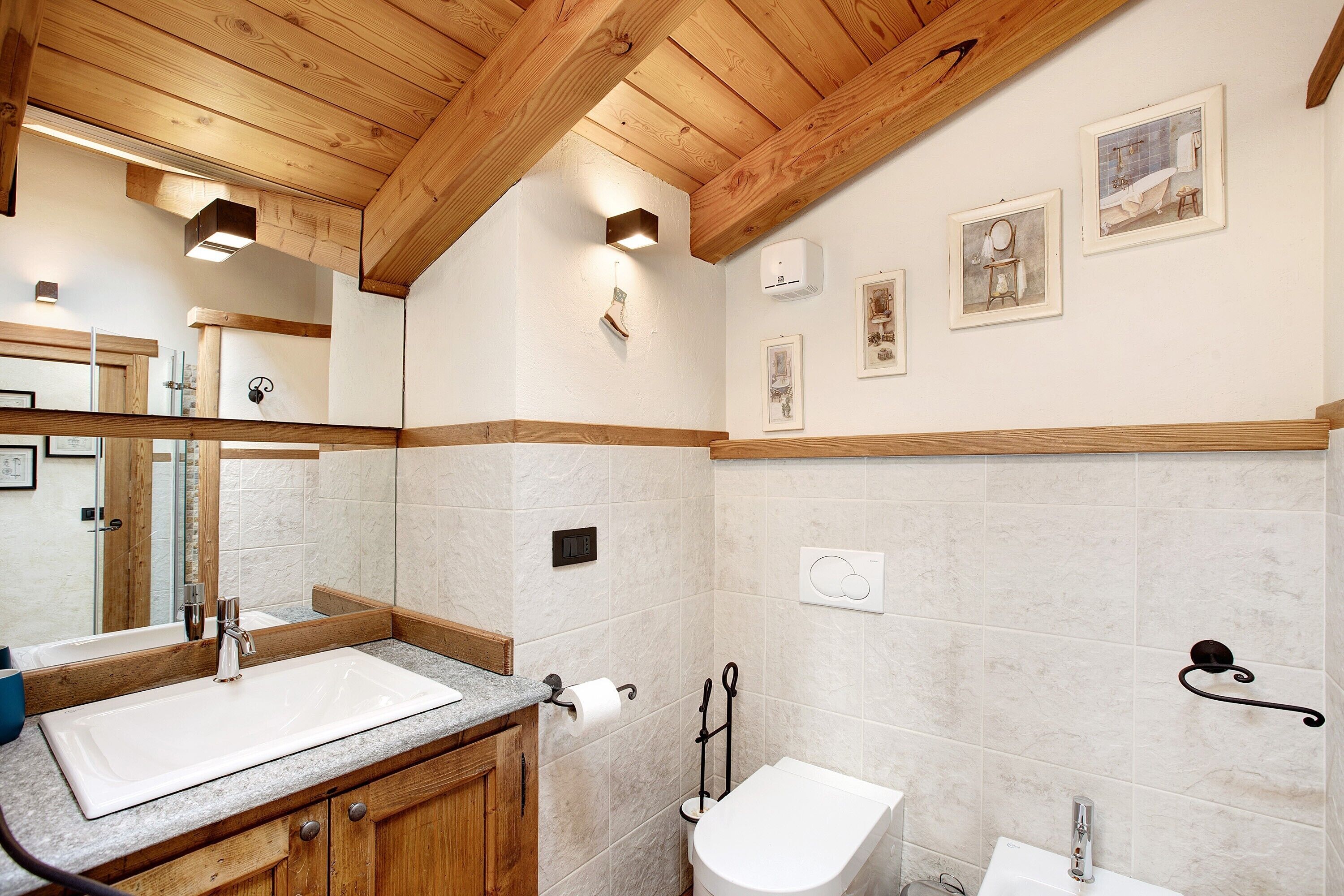 Un baño con techo de madera, un espejo grande, un lavamanos, un inodoro y cuadros enmarcados en la pared.