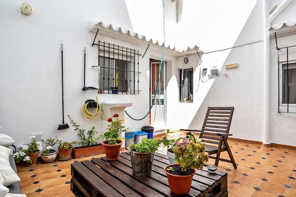 A patio with potted plants, a wooden table, a chair, and a white wall with a window and a door.