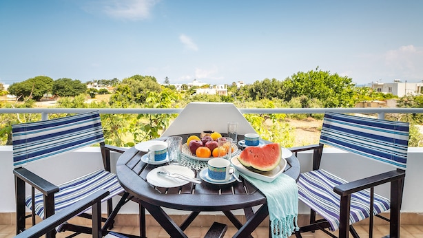 Ein Esstisch mit Stühlen, ein Grill und eine Obstplatte auf einem Balkon mit Blick auf Grünflächen und Gebäude.