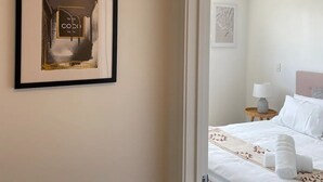 Hotellihuoneessa on sänky, yöpöytä, lamppu ja seinällä oleva kehystetty kuva, jossa on esillä hajuvesipullo.