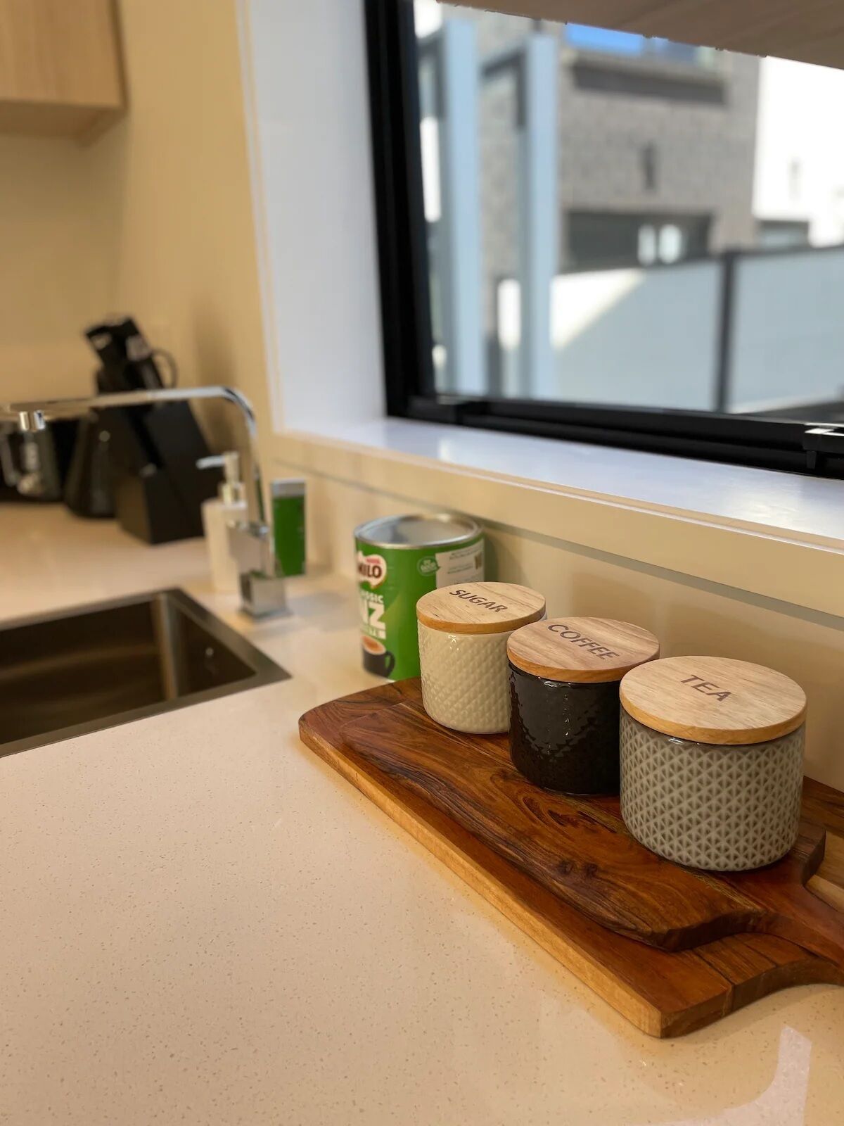 A kitchen counter with a wooden cutting board holding three containers labeled "SUGAR," "COFFEE," and "TEA." A can of "KILO" coffee is visible in the background.