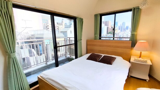 A hotel room with a large bed, a nightstand, a lamp, and a view of the city through large windows. - Minamiaoyama 1st (Tokyo)