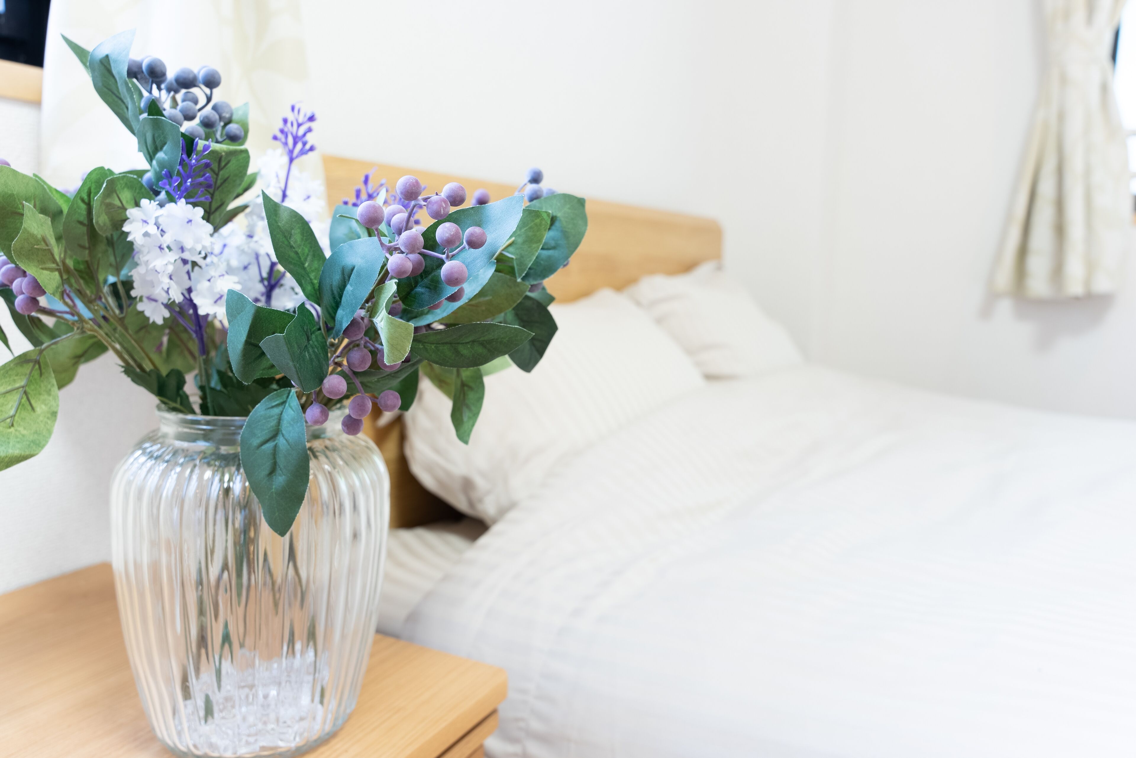 Un florero de vidrio transparente con un arreglo floral sobre una mesita de noche de madera al lado de una cama con ropa blanca.