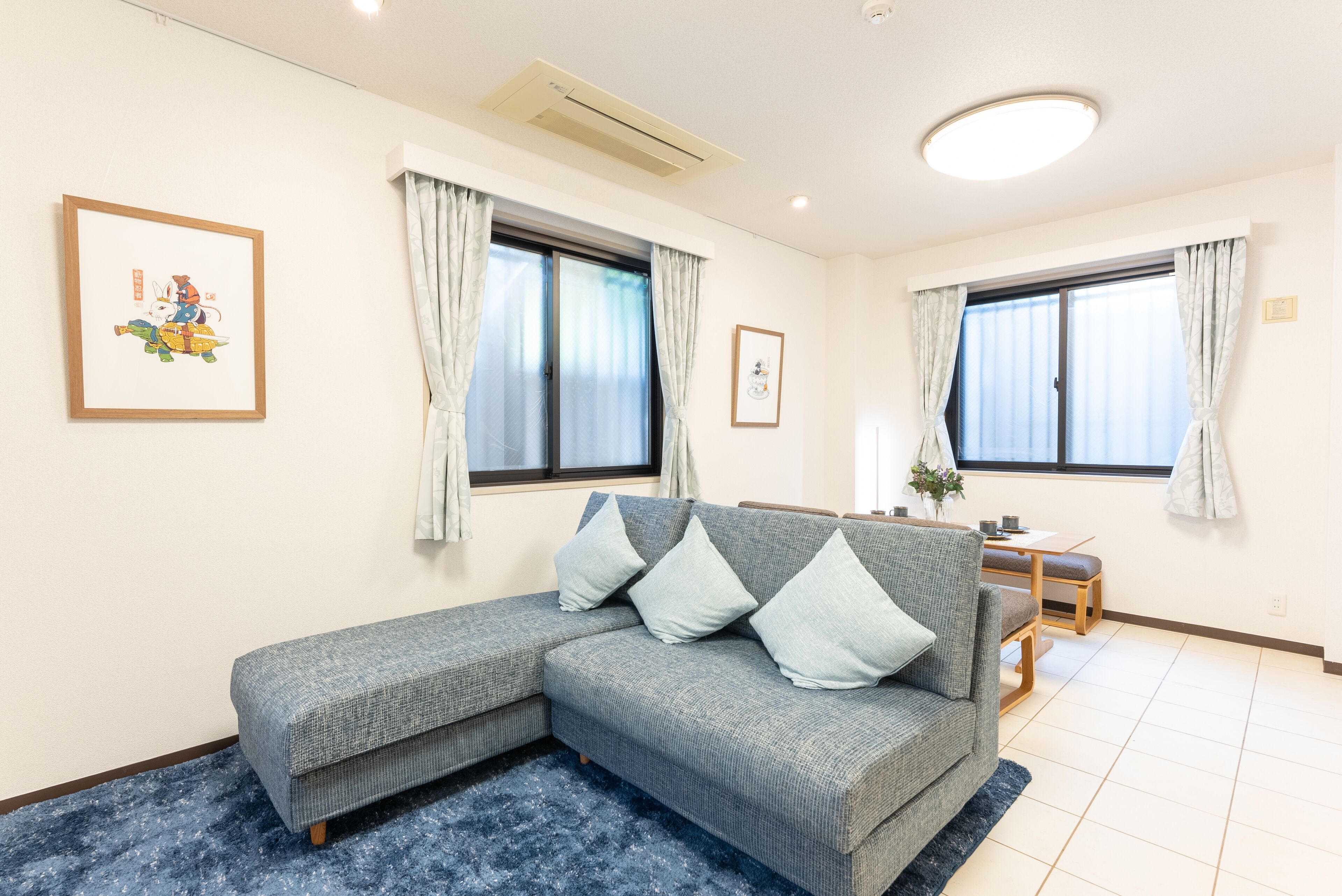 A living room with a grey sectional sofa, two windows with curtains, a small table with chairs, and a blue rug.