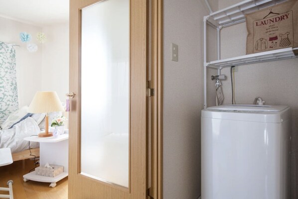 A bedroom with a bed, bedside table, lamp, and a laundry shelf.