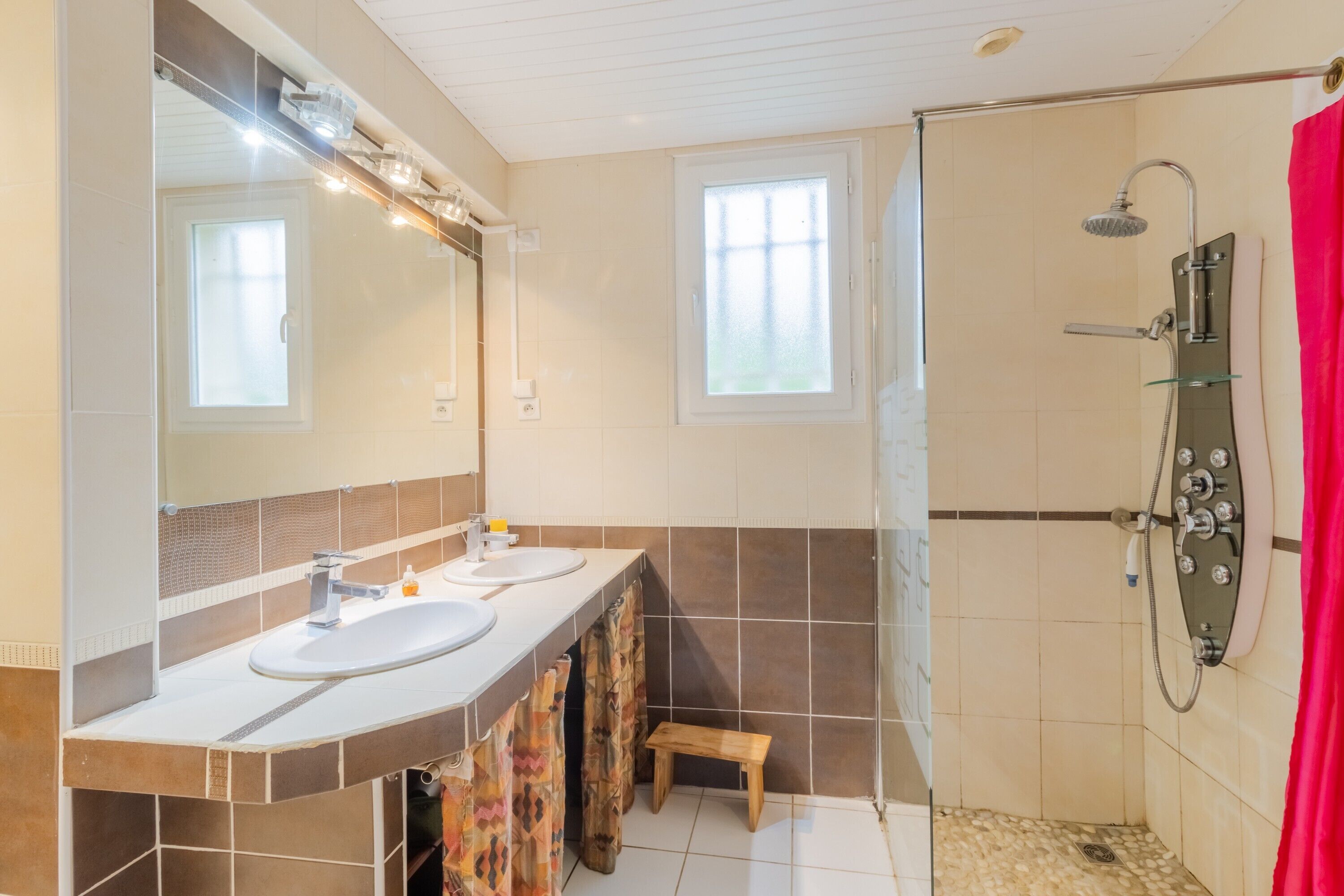 Un baño con dos lavamanos, ducha, un taburete pequeño y una ventana.