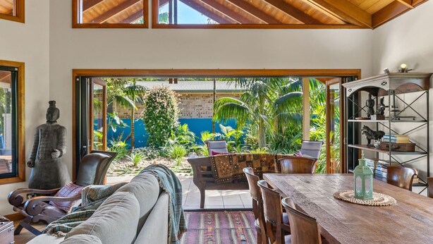 Un vaste espace de vie doté d’un plafond en bois, de grandes fenêtres, d’un canapé, d’une table à manger et offrant une vue sur un jardin luxuriant.