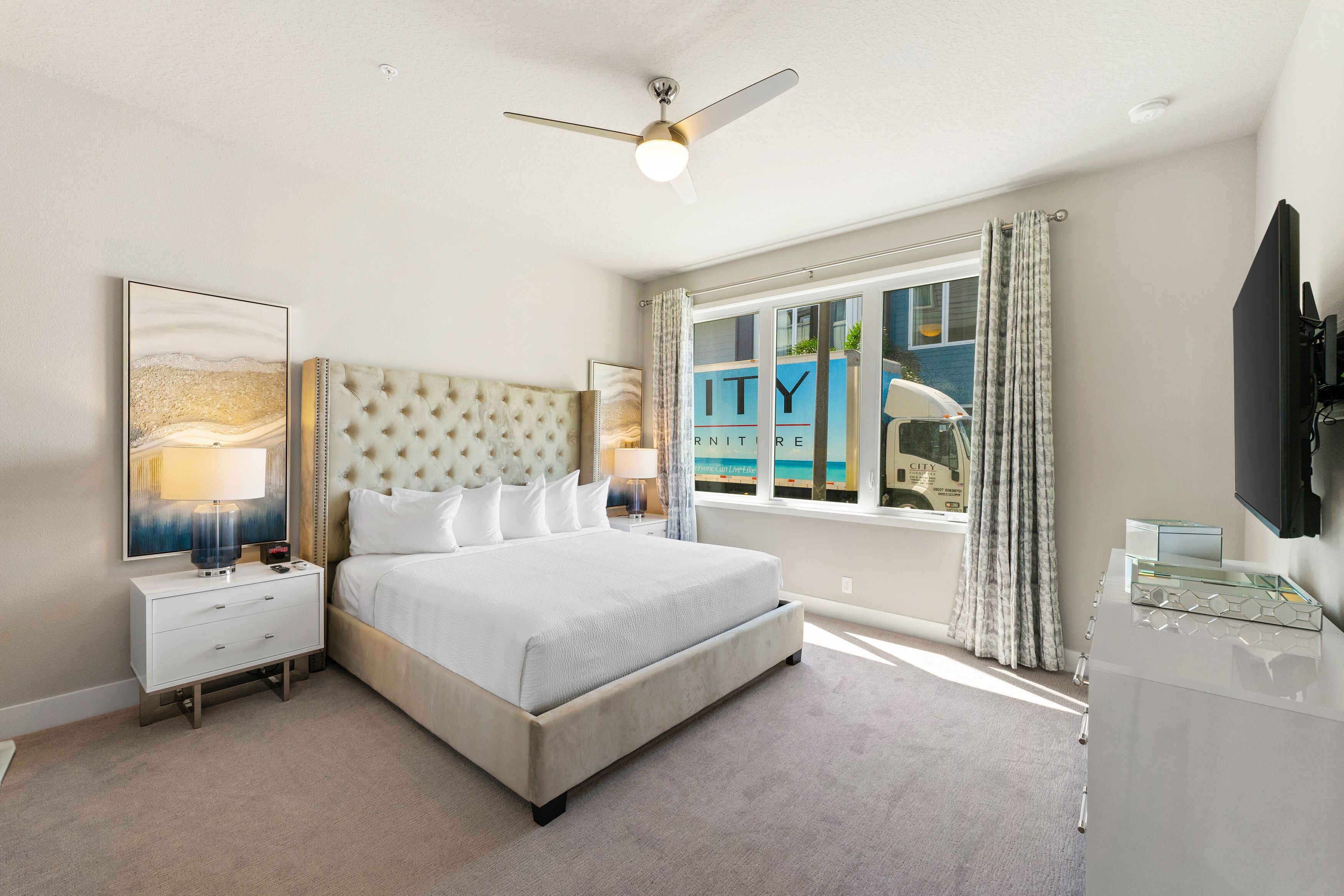 A modern hotel bedroom with a tufted headboard, a large bed, a nightstand with a lamp, a glass side table, and a flat-screen TV.