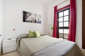 A bedroom with a bed, a nightstand, a lamp, a window with red curtains, and a cityscape painting on the wall.