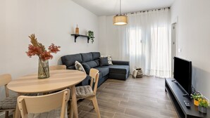 Een moderne woonkamer met een bank, een eettafel met stoelen, een televisie op een standaard en een hanglamp.