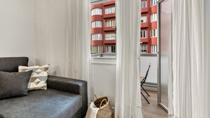 Een moderne woonkamer met een grijze bank, een gekleurd kussen, een geweven tas, en uitzicht op een rood gebouw door dunne gordijnen.