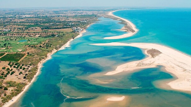 An aerial view of a coastal area with a sandy beach, clear blue waters, and a green landscape with scattered buildings. - Apartment "Sunset House Algarve" with Private Terrace & Wi-Fi (Faro)