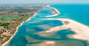 Luftaufnahme einer Küstenregion mit Sandstrand, klarem, blauem Wasser und einer grünen Landschaft mit vereinzelten Gebäuden.