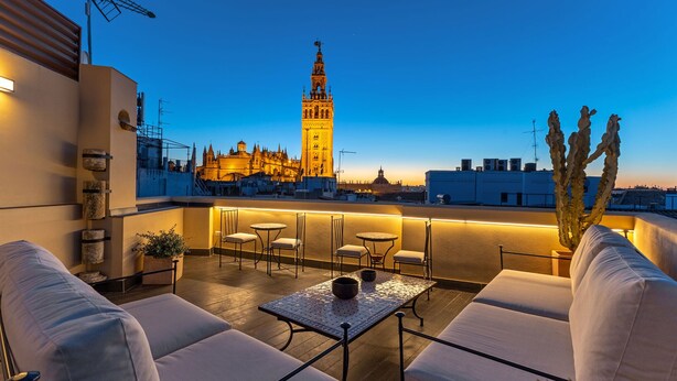Una terraza en la azotea con vistas a una torre histórica, amueblada con sofás y mesas blancos, y una planta de cactus.