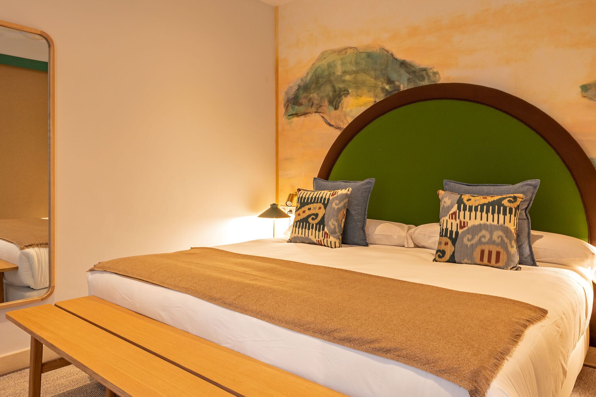 A neatly made bed with a green headboard, two patterned pillows, a brown throw blanket, and a wooden bench beside it.