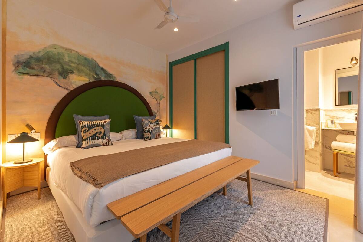 Un dormitorio moderno con una cama grande, un banco de madera, un televisor montado en la pared y un baño visible a través de una puerta abierta.