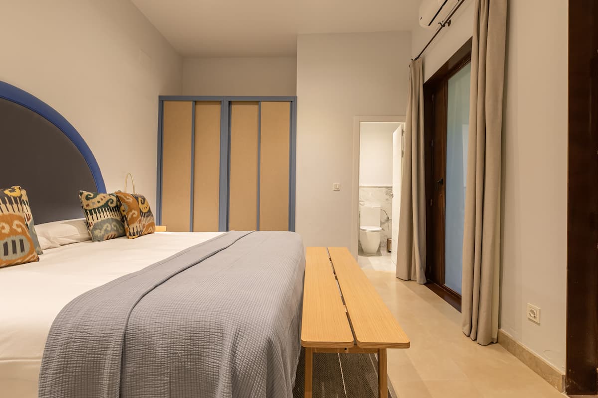 Un dormitorio con una cama, almohadas, un banco de madera, un armario y un baño visible al fondo.