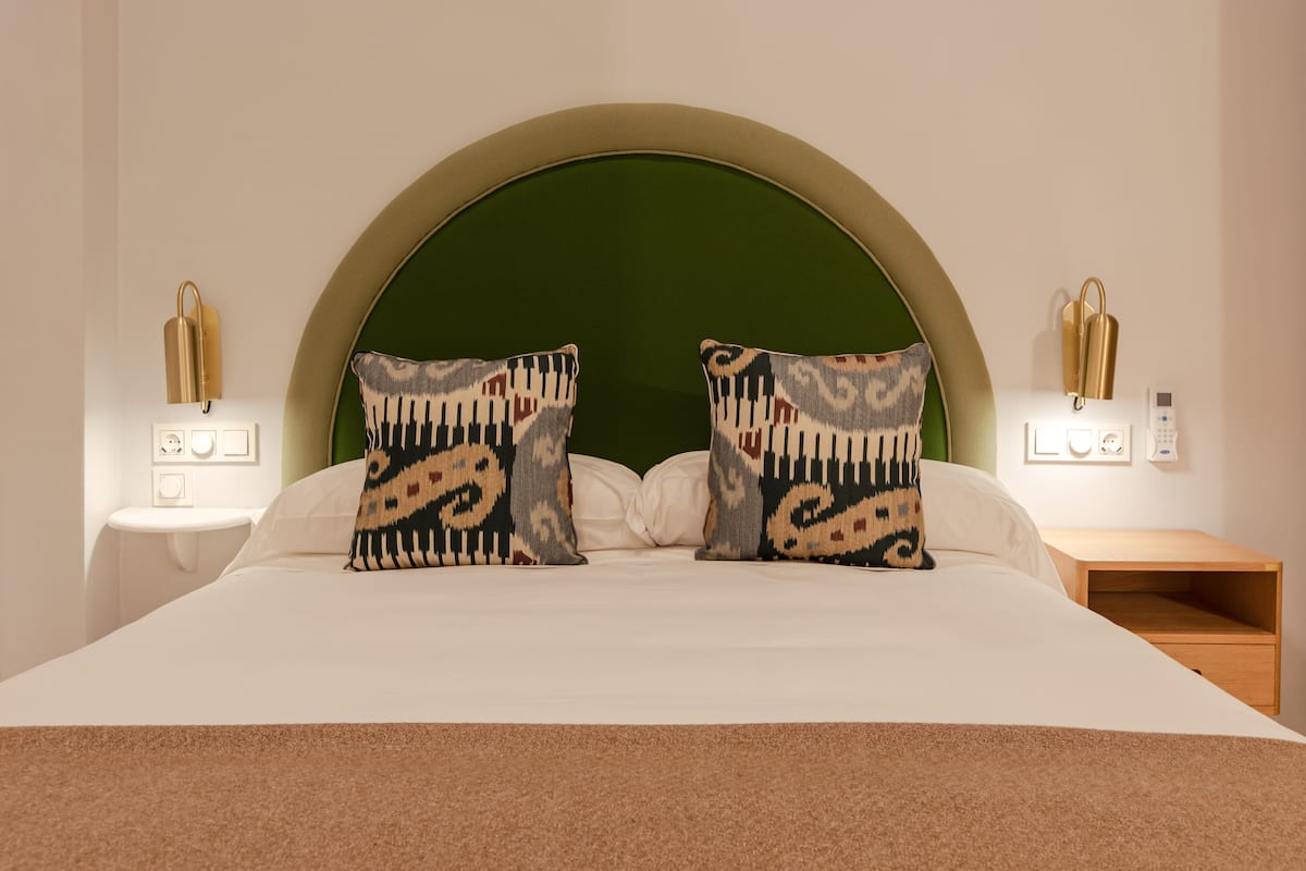 Una cama bien hecha con almohadas estampadas, un cabecero verde, mesitas de noche con lámparas y una mesita auxiliar.
