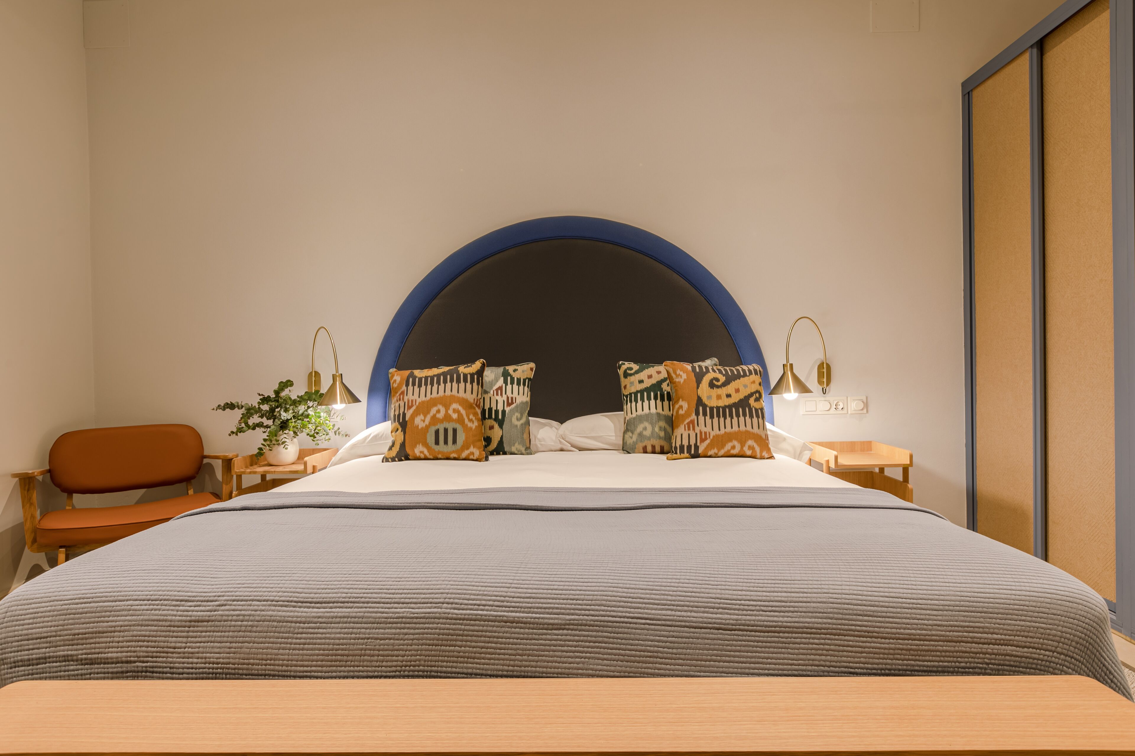 A modern bedroom with a large bed, a wooden bench, a small table with a plant, and a nightstand with a lamp.