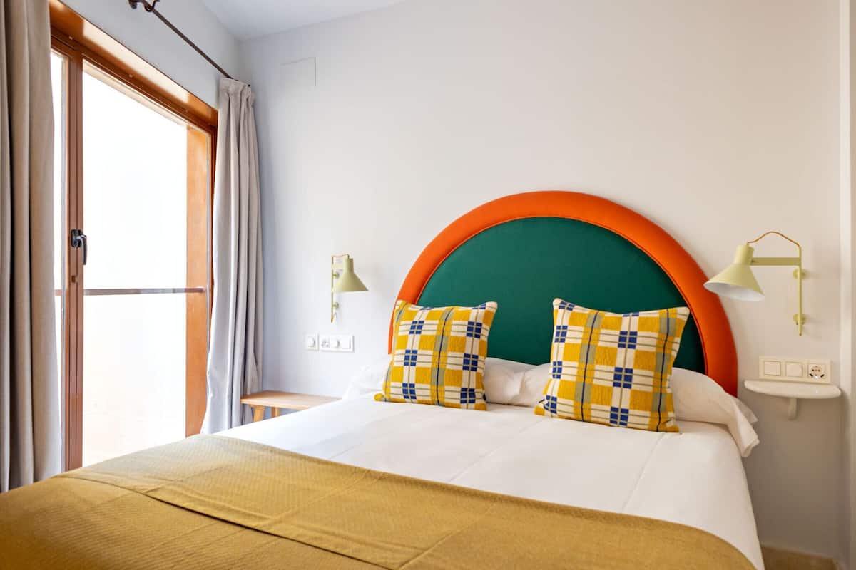 Habitación de hotel con una cama grande, dos almohadas, cabecero, lámparas de noche, ventana con cortinas y vistas al exterior.