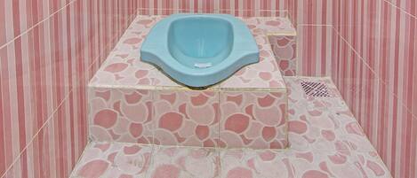 Un baño pequeño con baldosas rosas y blancas a rayas, un inodoro asiático azul y una lámpara de techo blanca.