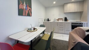 A compact kitchen with white cabinets, a built-in oven, and a coffee machine. There is a dining area with a white table and chairs, and a painting on the wall.
