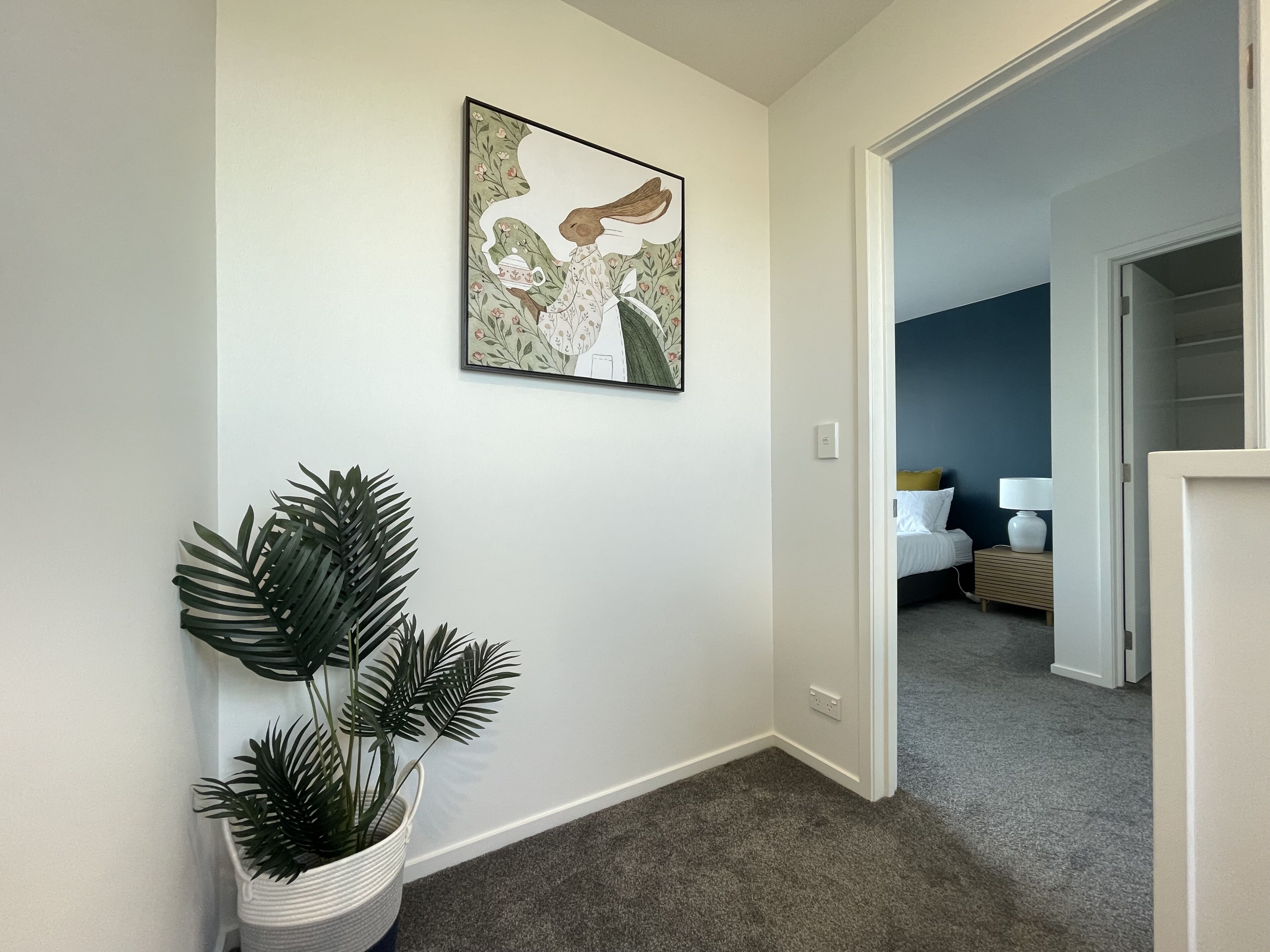 Ein Flur mit einem Blumentopf, einem gerahmten Kunstwerk und einem Blick in einen Schlafbereich mit Bett und Nachttisch.