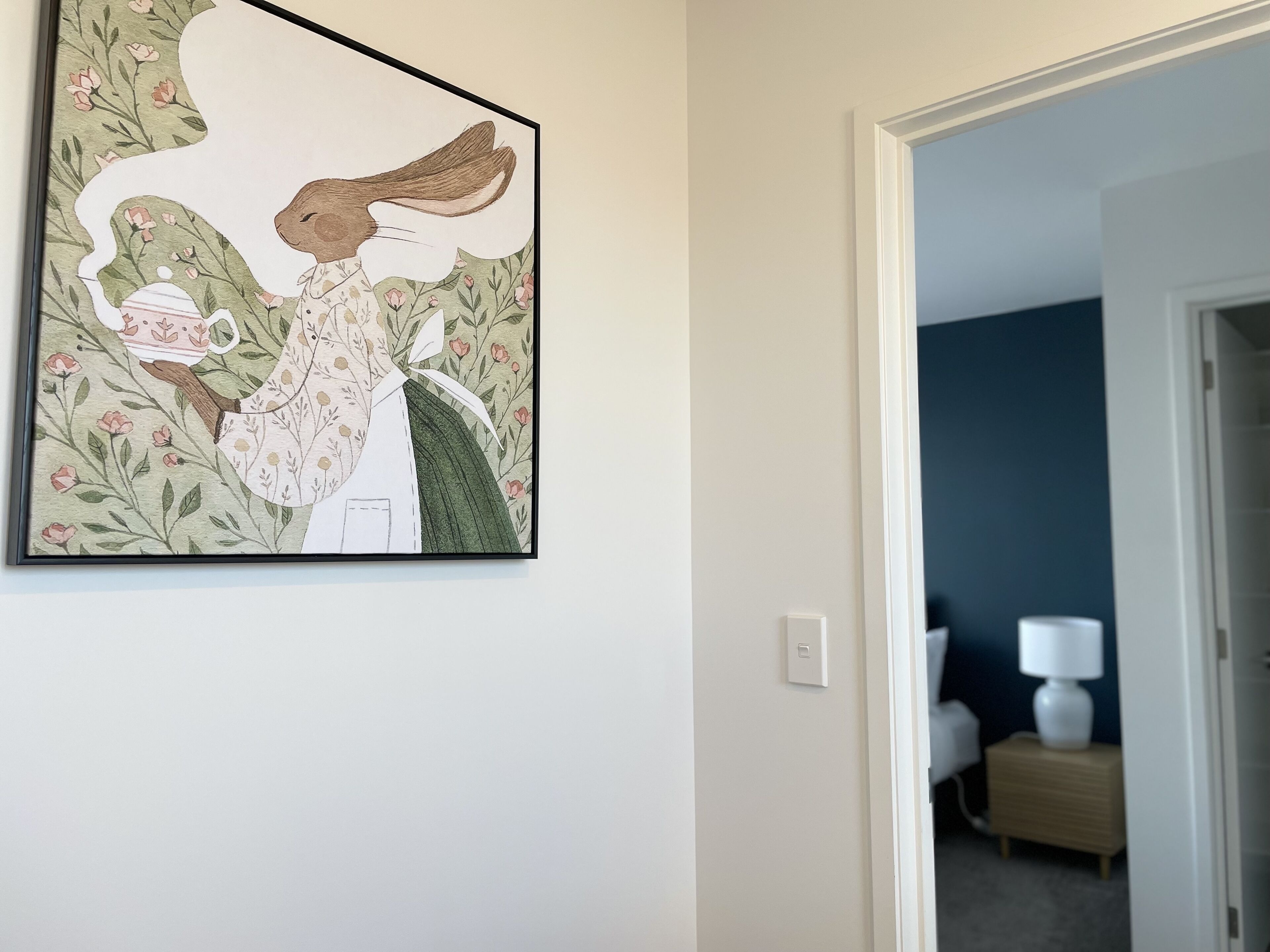 Une œuvre d’art encadrée accrochée au mur, une porte-fenêtre donnant sur une autre pièce et une table de chevet avec une lampe.