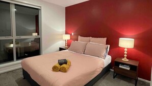 A modern bedroom with a large bed, two bedside tables with lamps, a red accent wall, and a sliding glass door leading to a balcony.