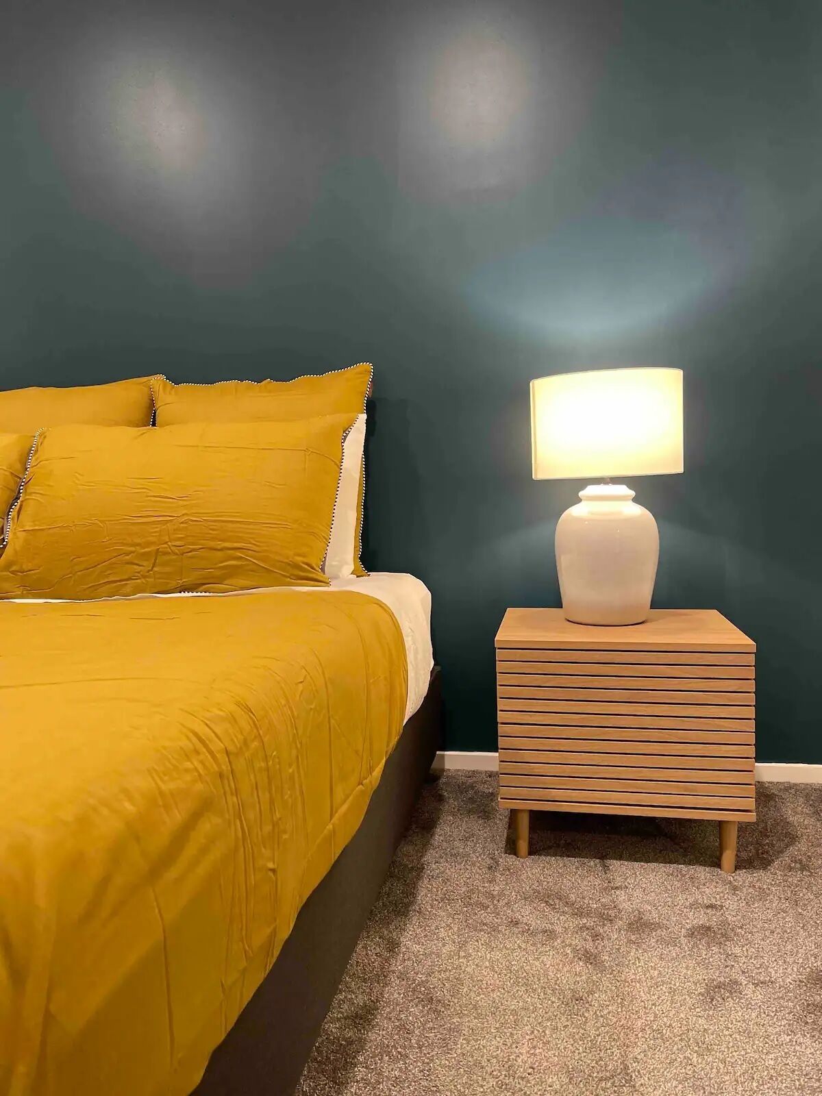 Ein Bett mit gelber Bettdecke und Kissen, eine weiße Nachttischlampe auf einem Holznachttisch und eine dunkel türkisfarbene Wand.