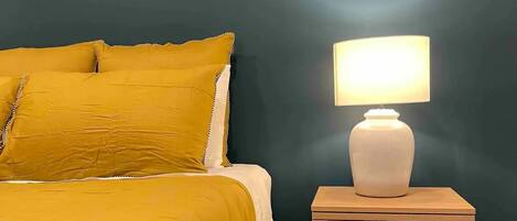 Ein Bett mit gelber Bettdecke und Kissen, eine weiße Nachttischlampe auf einem Holznachttisch und eine dunkel türkisfarbene Wand.