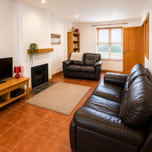 A living room with a fireplace, leather sofa, wooden bookshelf, and a television. - Kilkee Townhouse (Kilkee)