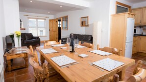A dining area with a wooden table set for six, surrounded by wooden chairs. A black leather sofa and a small wooden table with a vase of flowers are in the background. - Kilkee Townhouse (Kilkee)