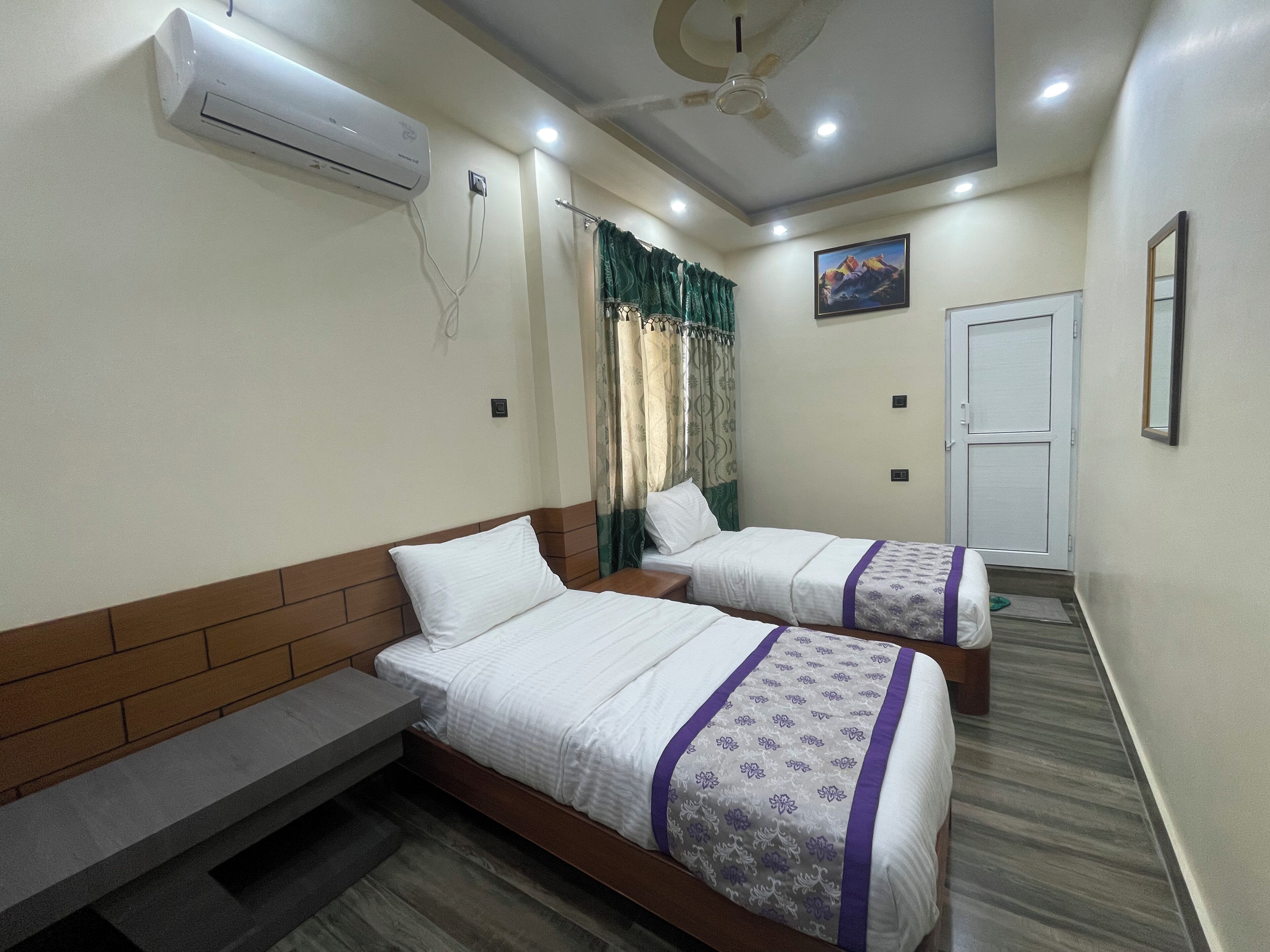 Habitación de hotel con dos camas, un banco, unidad de aire acondicionado, ventilador de techo, una puerta y un cuadro en la pared.