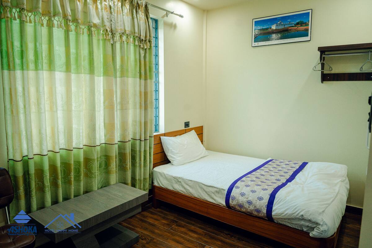 A single bed with a wooden frame, a white pillow, and a blue and white patterned blanket. A bench is placed at the foot of the bed, and a wooden shelf is mounted on the wall.