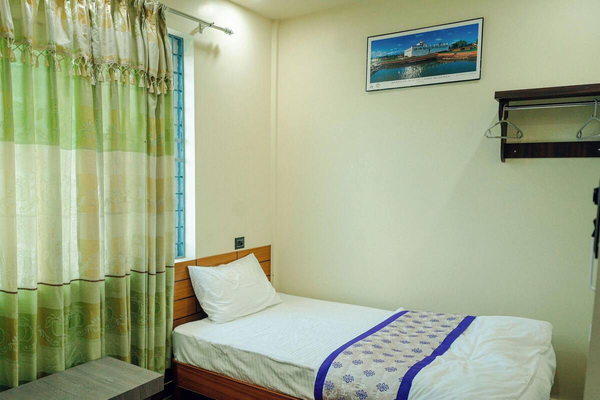 A single bed with a wooden headboard, a white pillow, and a blue patterned bedspread. A window with a blue curtain is visible, and a framed picture hangs on the wall.