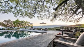 Zona junto a la piscina con sillones de descanso y sombrillas, rodeada de árboles y vistas a las colinas.