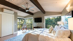 Un dormitorio con una cama grande, ventilador de techo, televisión y balcón con una silla y vistas a árboles.