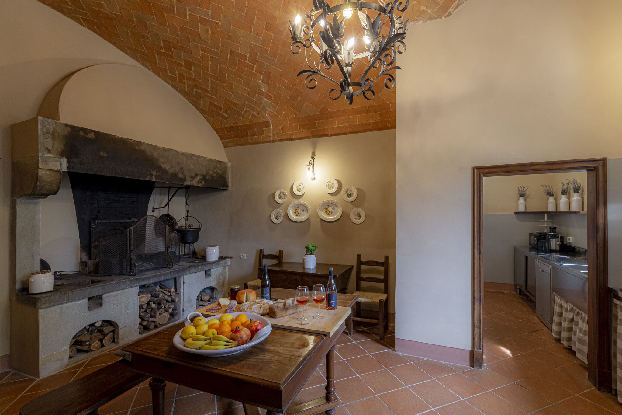 A rustic kitchen with a brick ceiling, a stone fireplace, and a wooden dining table set with fruits, wine, and bread.