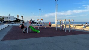 Une aire de jeux avec un toboggan vert et des structures d’escalade, située près d’une plage où le ciel est dégagé.