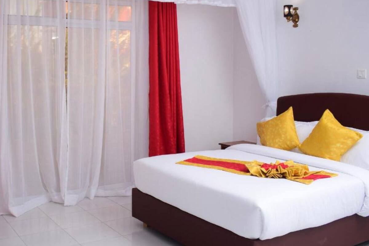 A hotel room with a bed, white curtains, a red curtain, and a wall-mounted light fixture.