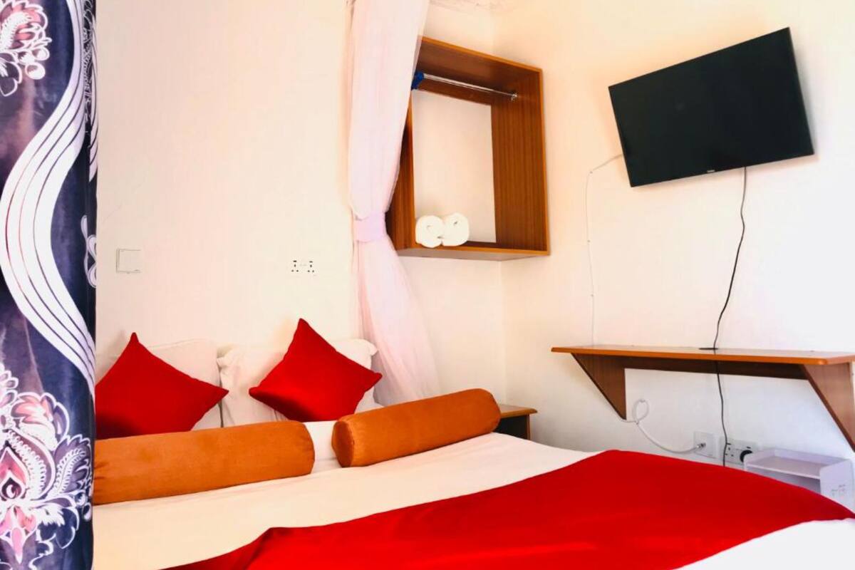 A bedroom with a bed, red and orange pillows, a wooden bedside table, a mirror, and a TV mounted on the wall.